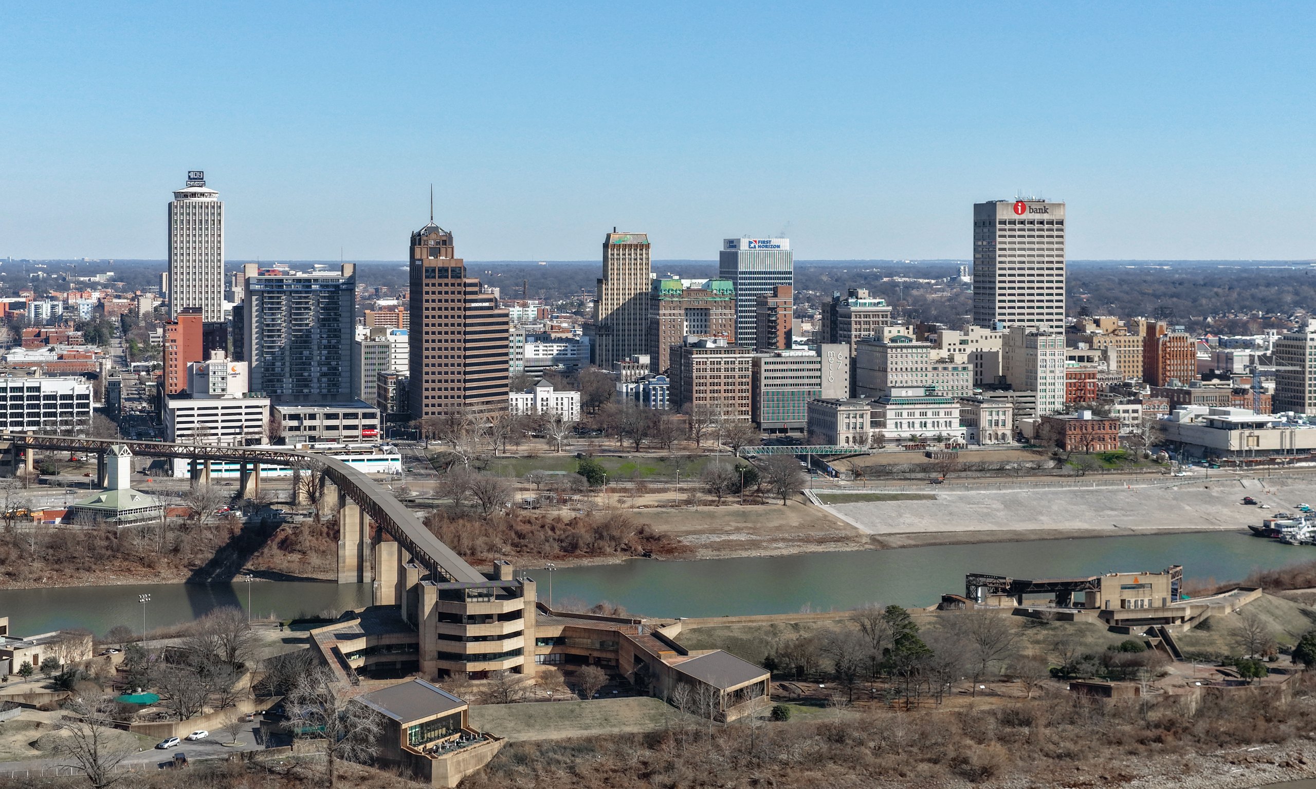 The Memphis skyline along the Mississippi River &mdash; the downtown towers standing on the bluff above the brown water, with the arched bridge crossing to Arkansas beyond.