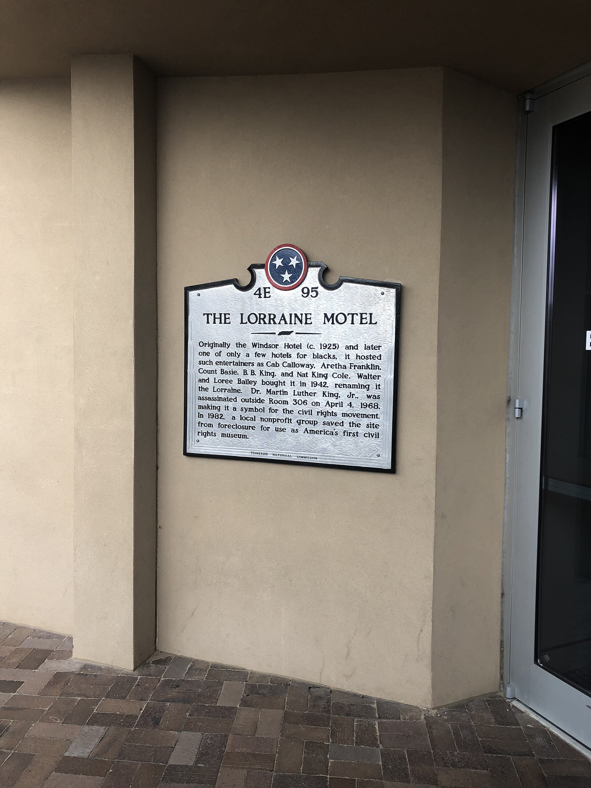 Silver Tennessee historical marker for The Lorraine Motel mounted on a beige stucco wall beside a glass door
