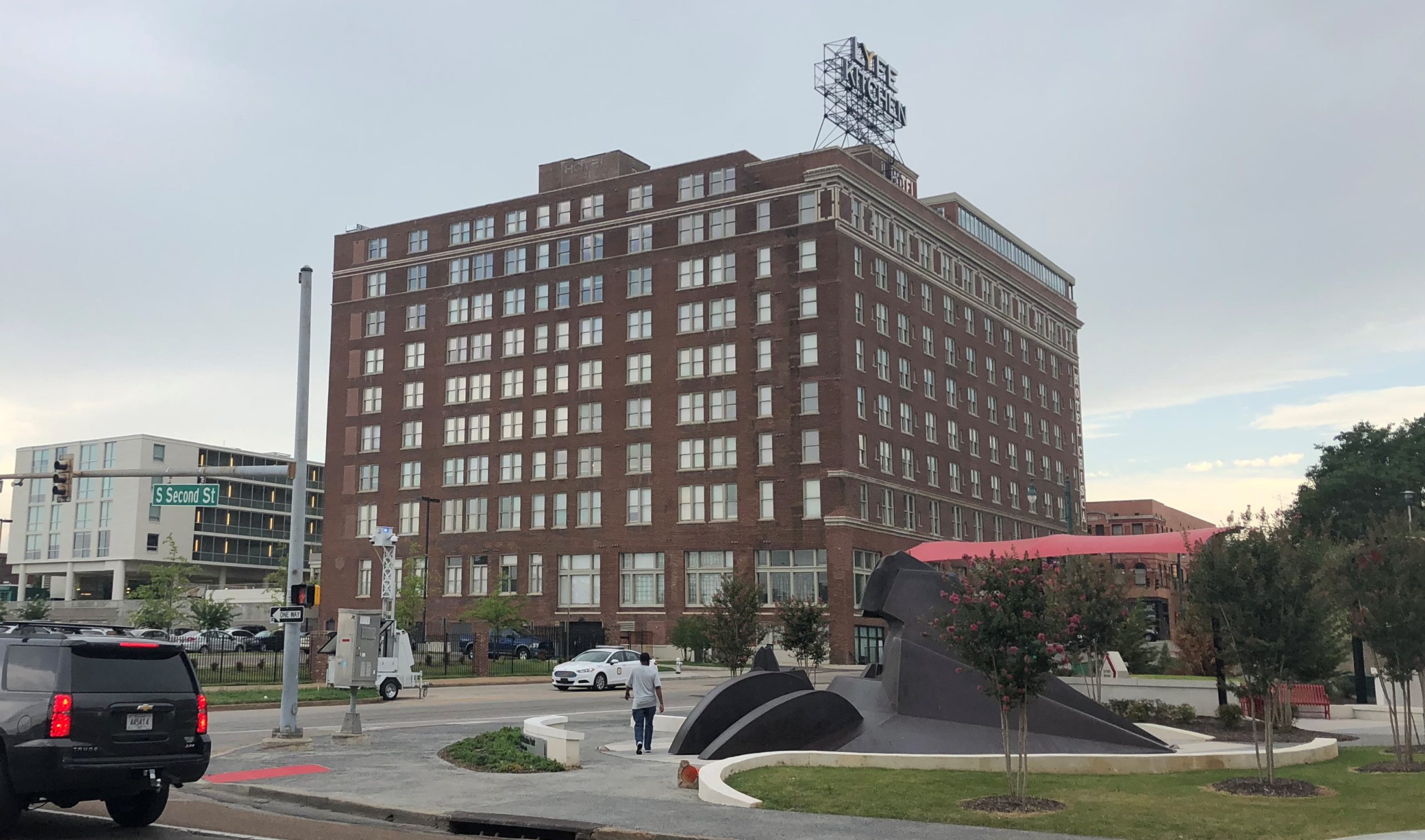 Tall brick historic hotel with a rooftop sign, seen across a street corner with an SUV, a pedestrian and a dark angular sculpture