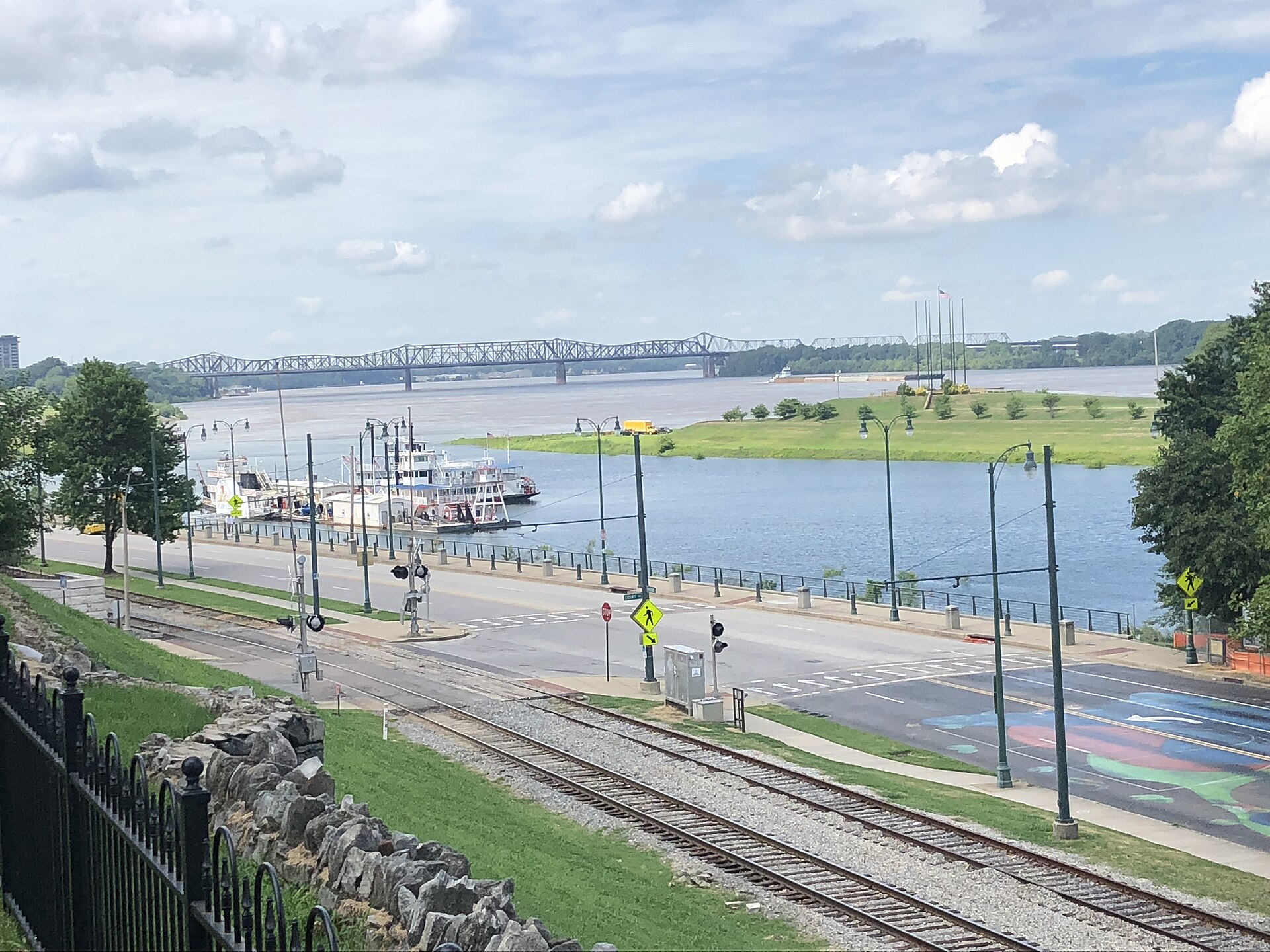 Riverfront view over railway tracks and a road to a moored riverboat, grassy island and steel truss bridge across the Mississippi