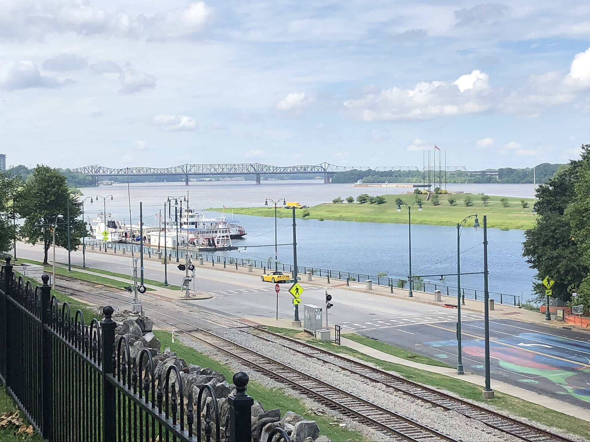 Riverfront road and railway tracks beside moored riverboats, with a steel truss bridge crossing the wide river beyond
