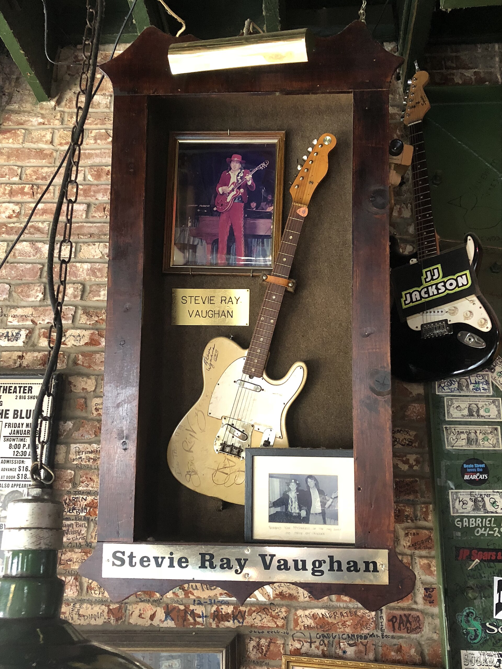Wooden display case on a graffiti-covered brick wall holding a signed guitar, framed photos and a Stevie Ray Vaughan plaque