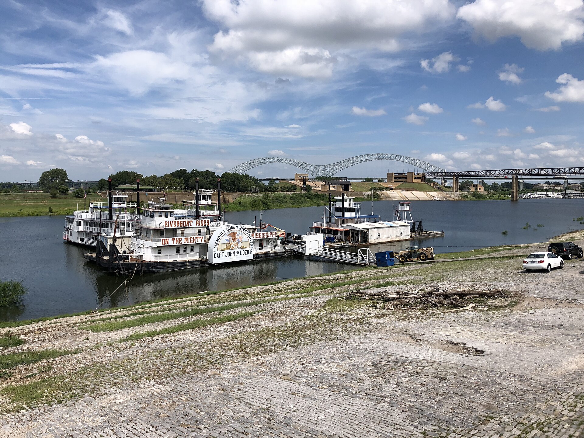 Paddlewheel riverboats moored along a cobblestone Mississippi riverbank in Memphis, with the arched Hernando de Soto Bridge beyond