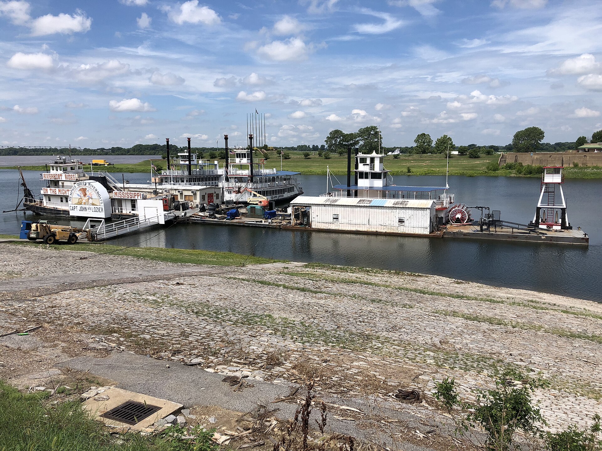Paddlewheel riverboats and barges moored along a cobblestone river landing under a partly cloudy sky